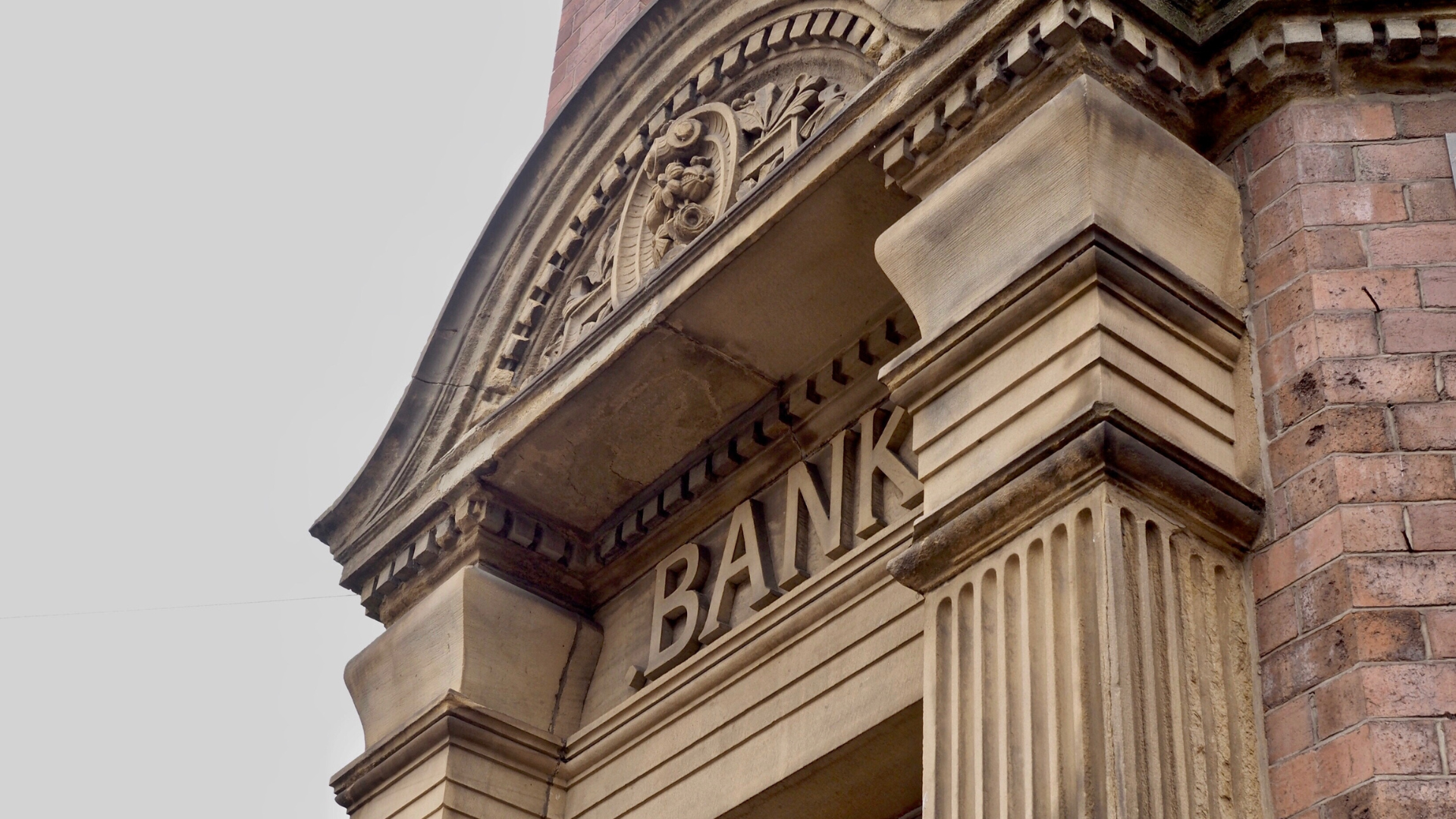 Close-up view of a classical bank façade showing a carved stone pediment and molding above an entrance with the word ‘BANK’ in large carved letters, a fluted column at right, red brickwork on the far right, and a pale overcast sky in the background
