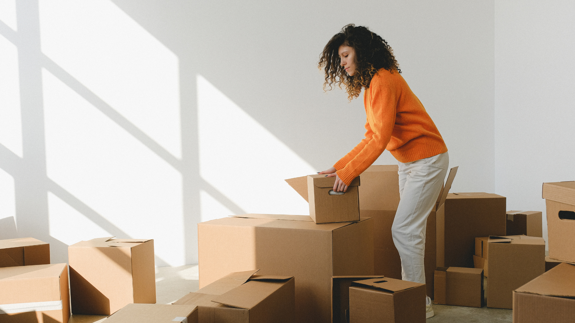 A lady packing her A curly-haired person in an orange sweater and light trousers leans forward to place or lift a small cardboard box among medium and large moving boxes, in a bright, minimalist room illuminated by sunlight from a window casting grid-like shadows on a plain light wall