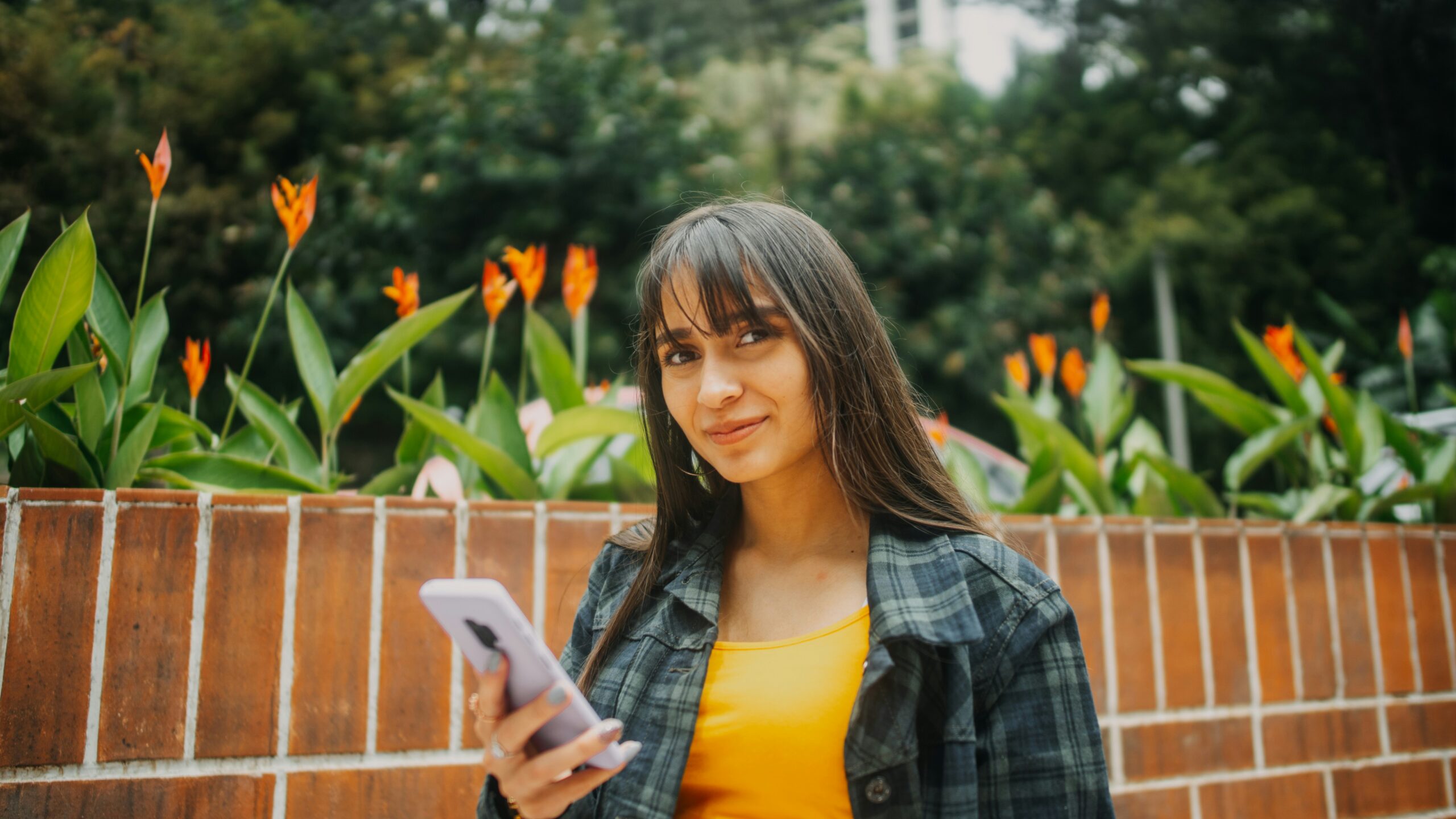 Student with long hair budgeting for uni, walking past a wall with flowers and a phone in her hand