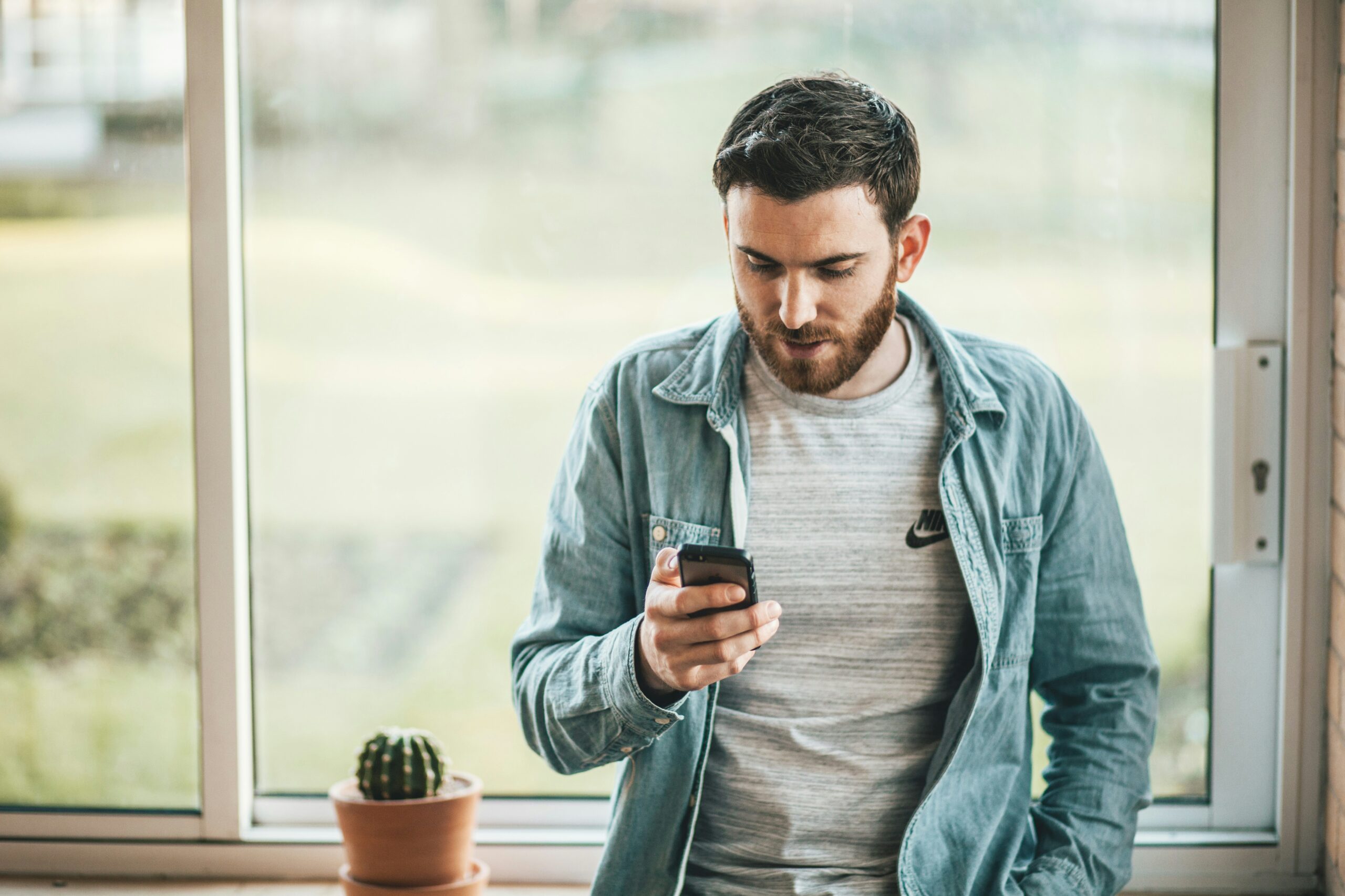 Young man standing by a window looking at a smartphone in his hand exploring save money apps