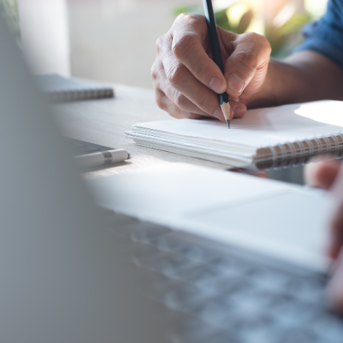 Someone writing with a pencil while working on a laptop