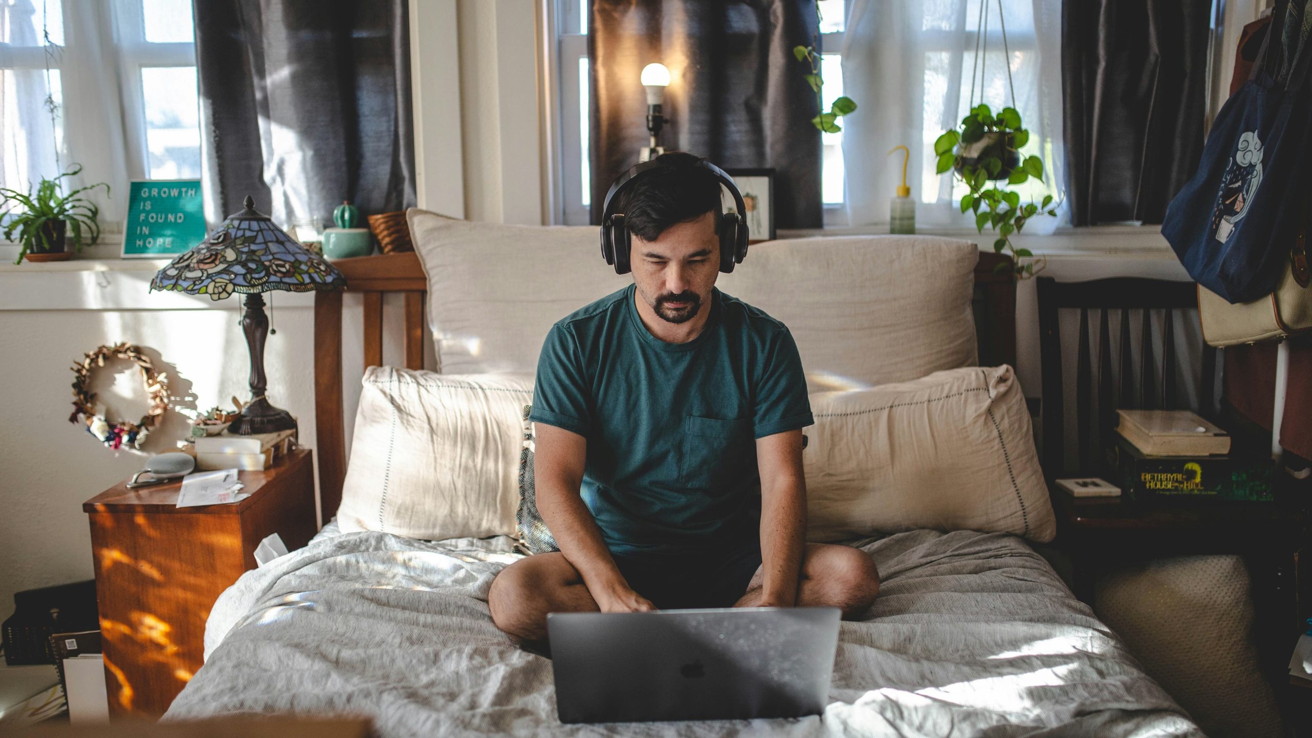 A young man sat on a bed wearing headphones and working on a laptop
