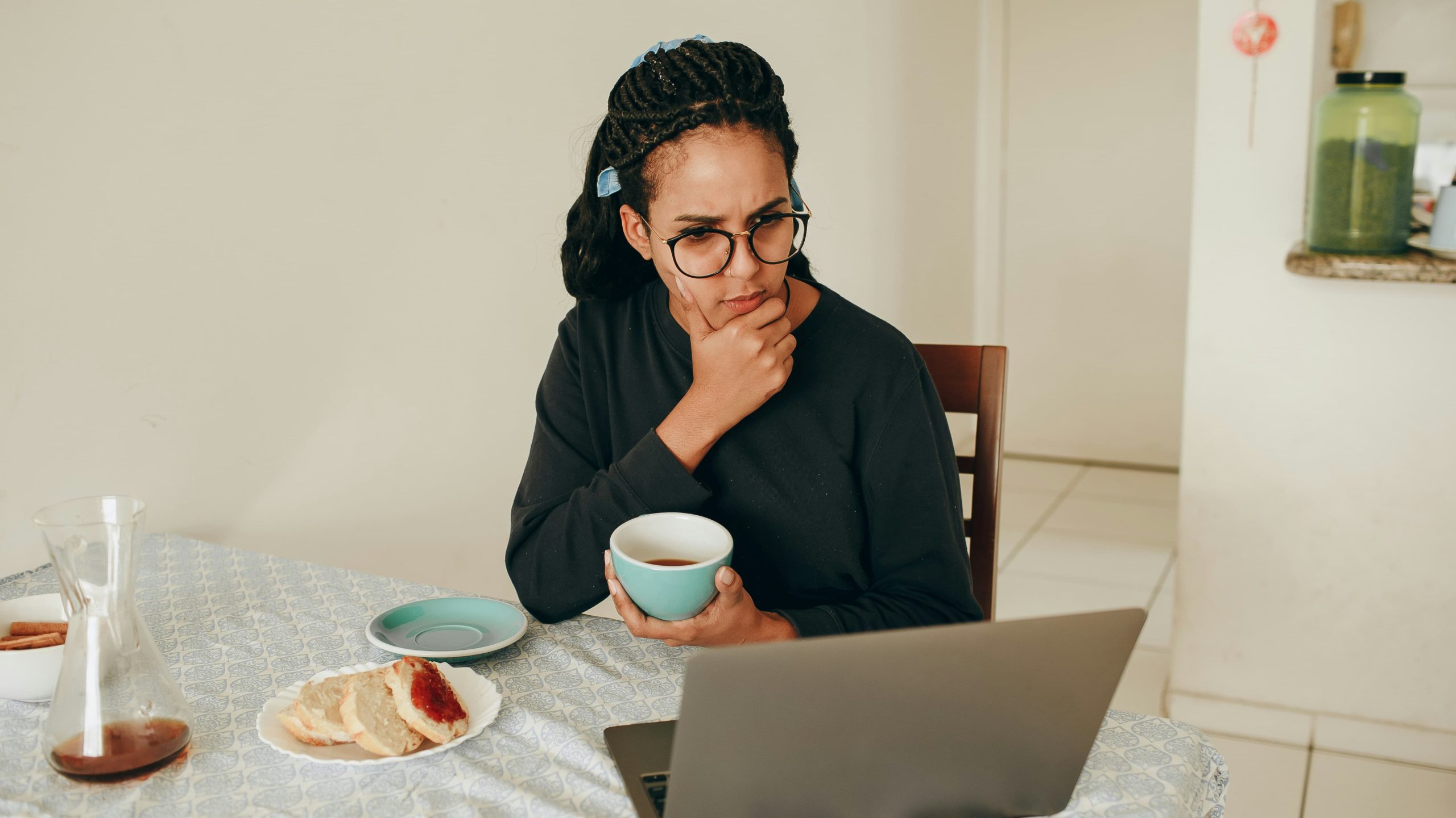 Young woman sat at her kitchen table with a cup of tea and snack and looking at her laptop