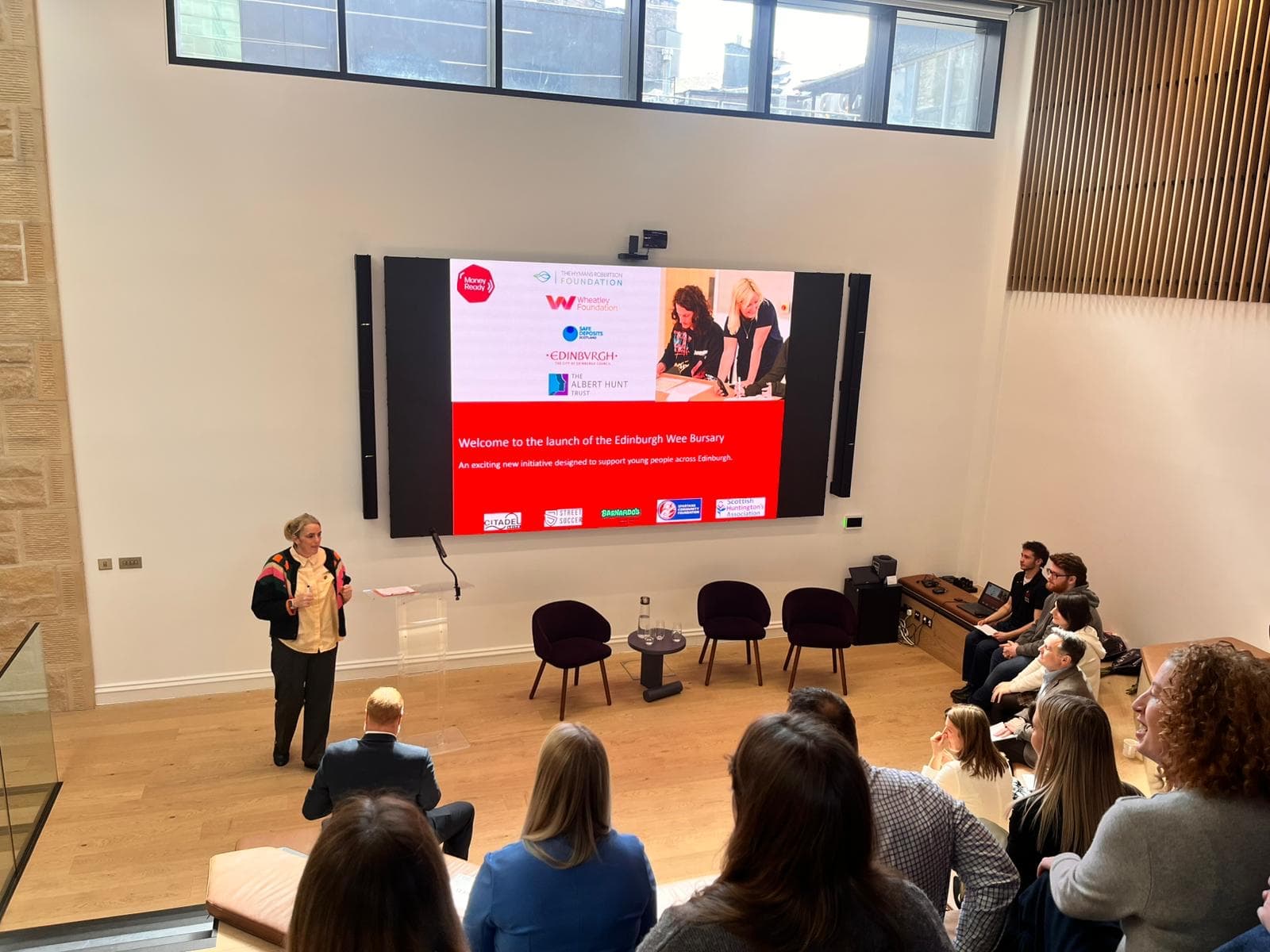 Helen Foster from Money Ready stands next to a screen presenting the Wee Bursary to a room of guests