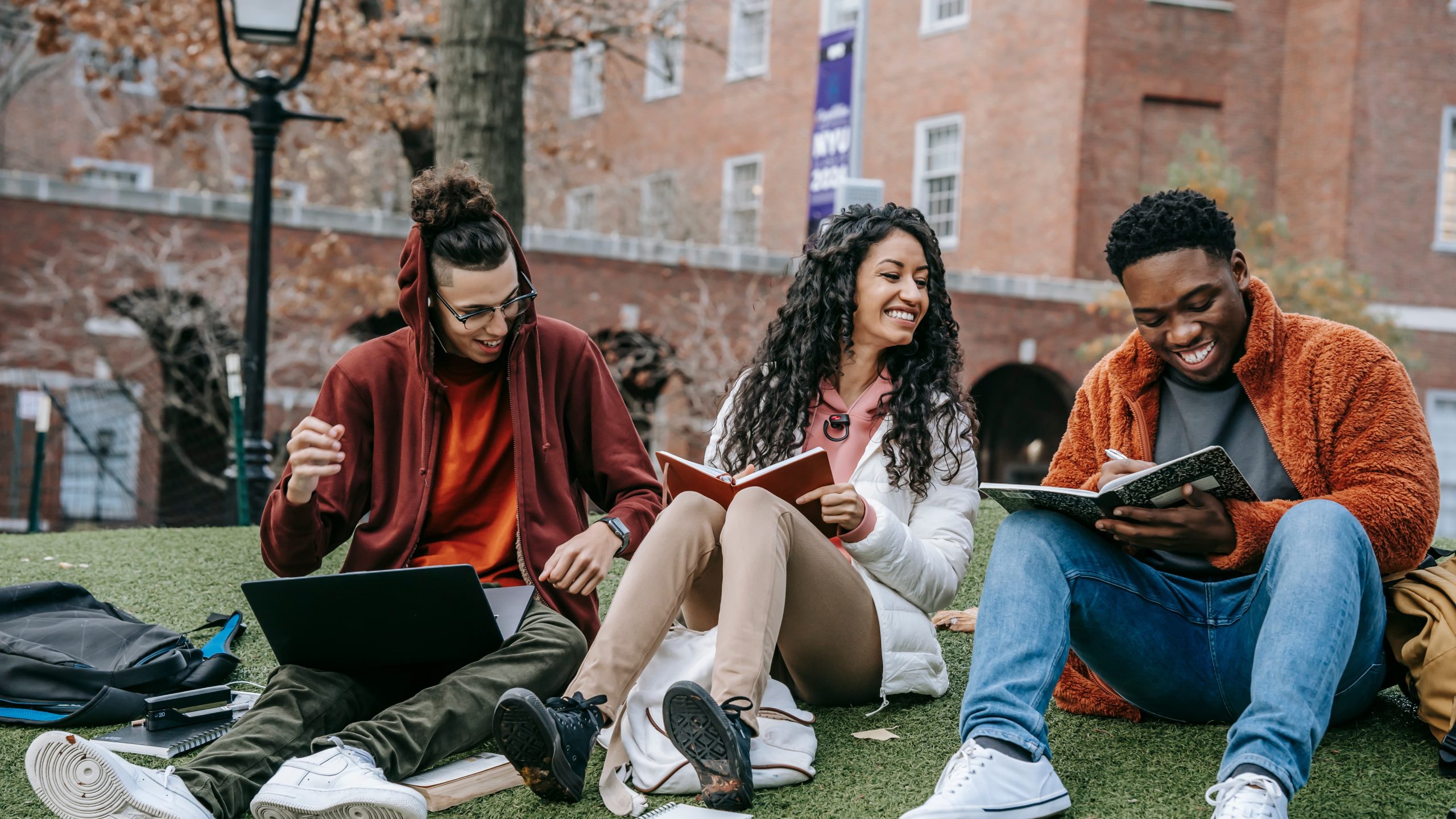 Three university students sitting together on artificial turf outdoors, laughing and studying. One young man in a maroon hoodie gestures while working on a laptop, a young woman in a white puffer jacket smiles while holding an open notebook, and another young man in an orange fleece jacket writes in a spiral notebook. Notebooks, backpacks, and a laptop are scattered around them on the grass, with a brick campus building visible in the background.