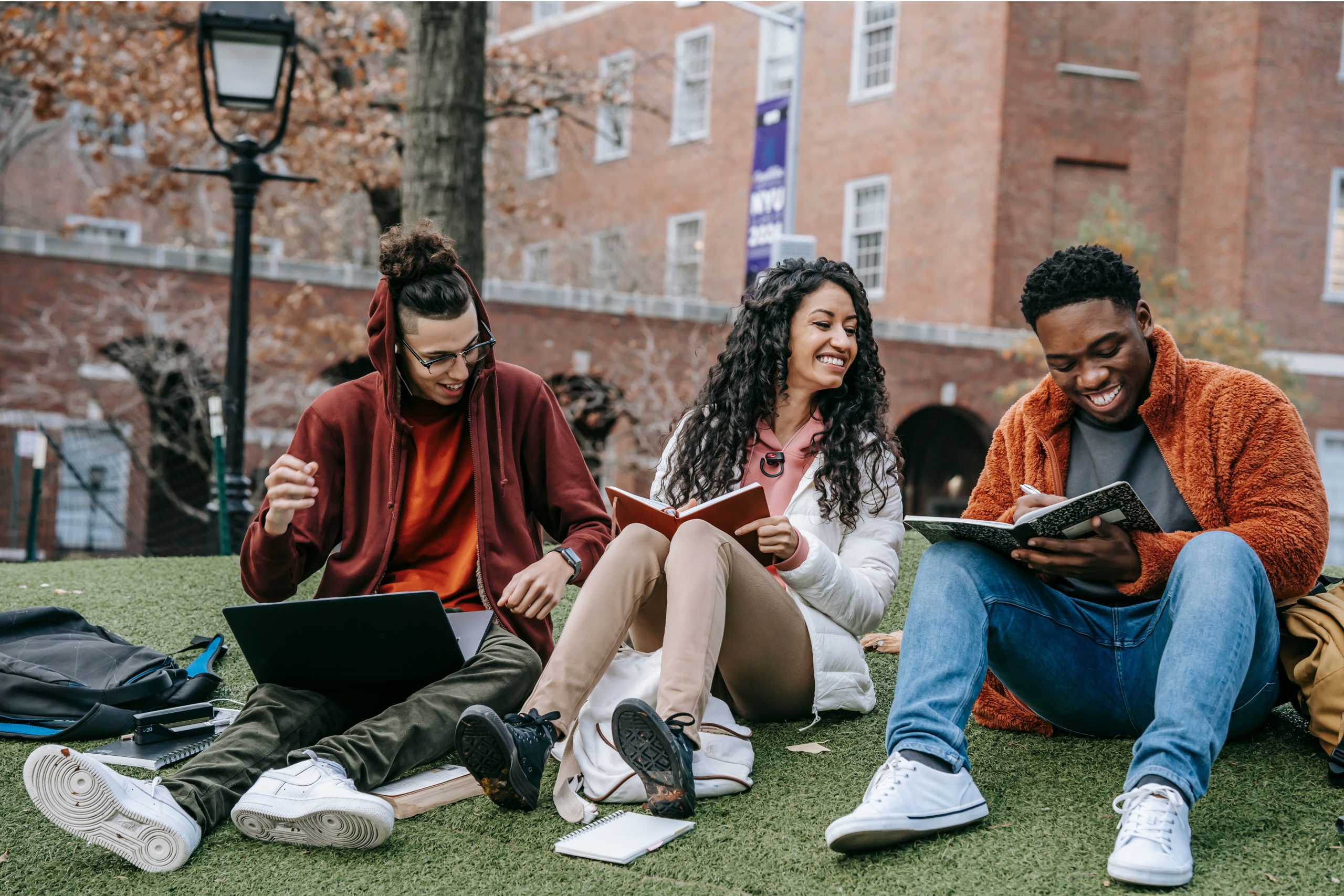 Three university students sitting together on artificial turf outdoors, laughing and studying. One young man in a maroon hoodie gestures while working on a laptop, a young woman in a white puffer jacket smiles while holding an open notebook, and another young man in an orange fleece jacket writes in a spiral notebook. Notebooks, backpacks, and a laptop are scattered around them on the grass, with a brick campus building visible in the background.