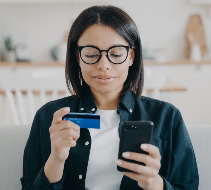 Person reading their phone and a bank card. They have dark straight hair and wear glasses.