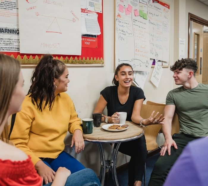 Four young people smiling, talking to eachother round a table with biscuits and tea.