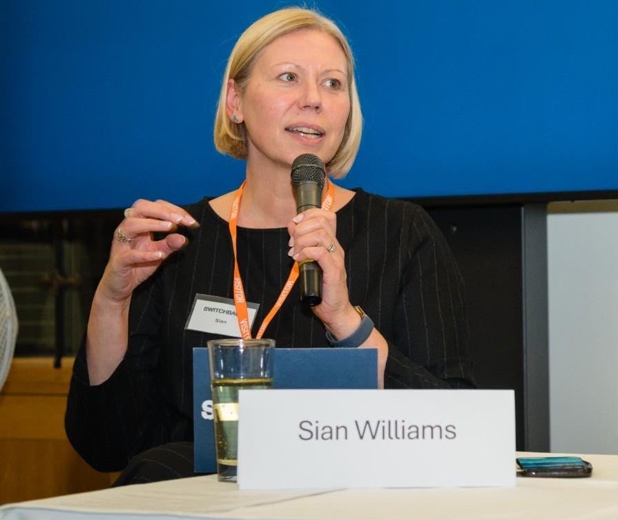 Sian Williams speaking at an event with a microphone. Sian has short blonde hair and wears a black top.